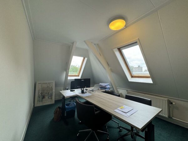 Kleine kantoorruimte met schuin dak, twee dakramen, een bureau met stoelen en stapels papier.